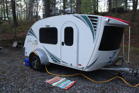Cozy white teardrop RV rental set up at a wooded campsite, with a yellow utility hose and a colorful rug on the gravel.