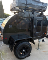 Black teardrop RV rental with rooftop tent, side hatch, and rugged all-terrain tires, ready for adventure.