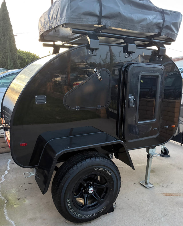 Black teardrop RV rental with rooftop tent, side hatch, and rugged all-terrain tires, ready for adventure.