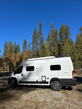 An RV rental, a white camper van, is parked in a serene forest campground with tall trees and autumn leaves under a blue sky.