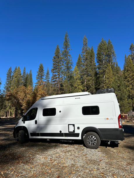 An RV rental, a white camper van, is parked in a serene forest campground with tall trees and autumn leaves under a blue sky.