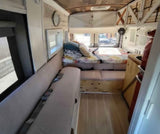 RV rental interior showcasing a cozy bed, long bench seat with seatbelts, wooden floor & ceiling, and white storage cabinets.
