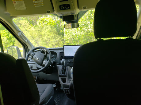 Interior view of an RV rental: front cabin with dashboard, large screen, steering wheel, and trees visible outside.
