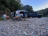 A couple enjoys a campfire beside their RV rental and Jeep on a rocky riverbed, perfect for an outdoor adventure.