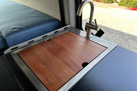 Inside an **rv rental**: a compact kitchen features a stainless steel faucet, sink with a Ruvati cutting board, and blue bed.