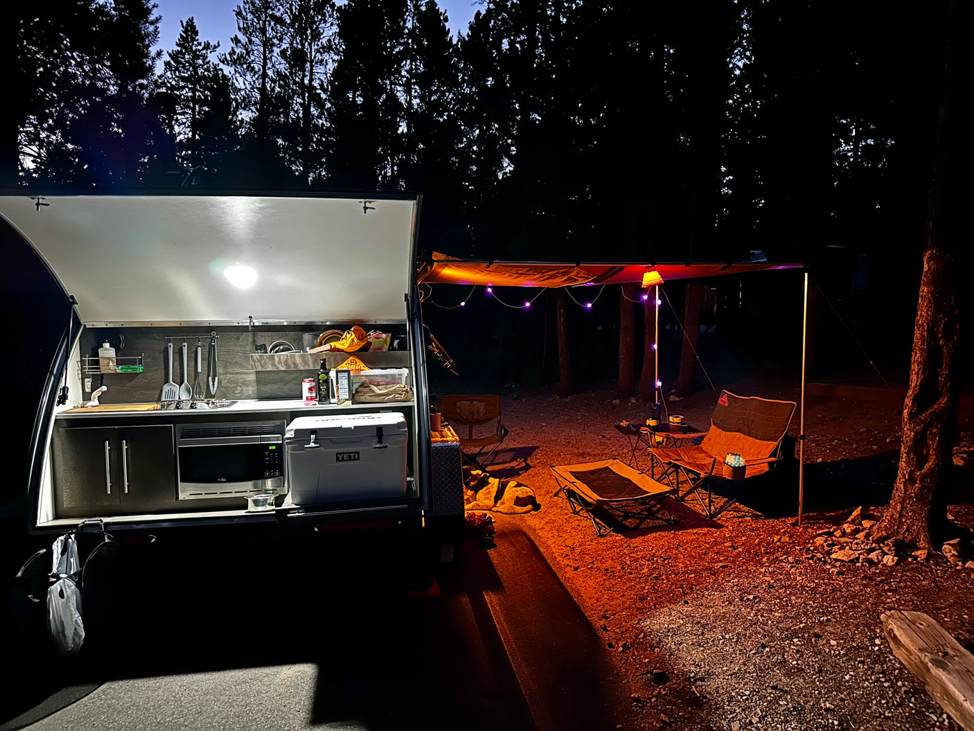 Night at a cozy RV rental campsite. Illuminated teardrop camper kitchen, string lights on awning, chairs, and a dog resting.