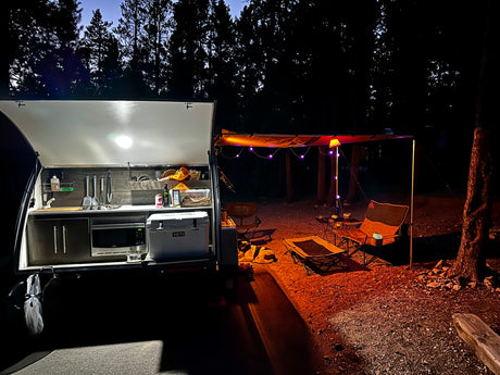 Night at a cozy RV rental campsite. Illuminated teardrop camper kitchen, string lights on awning, chairs, and a dog resting.