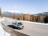 Silver RV rental van by WanderVans on a snowy mountain road. Pine trees and snow-capped peaks frame this winter travel scene.