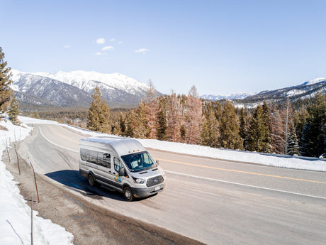 Silver RV rental van by WanderVans on a snowy mountain road. Pine trees and snow-capped peaks frame this winter travel scene.