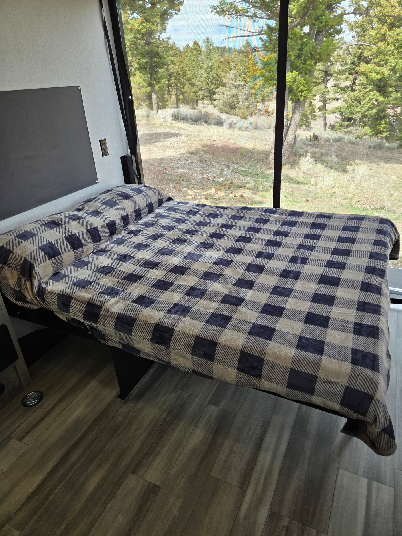 This RV rental features a cozy bed with a blue and beige plaid blanket, positioned by a large window showing a green forest view.