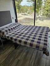 This RV rental features a cozy bed with a blue and beige plaid blanket, positioned by a large window showing a green forest view.