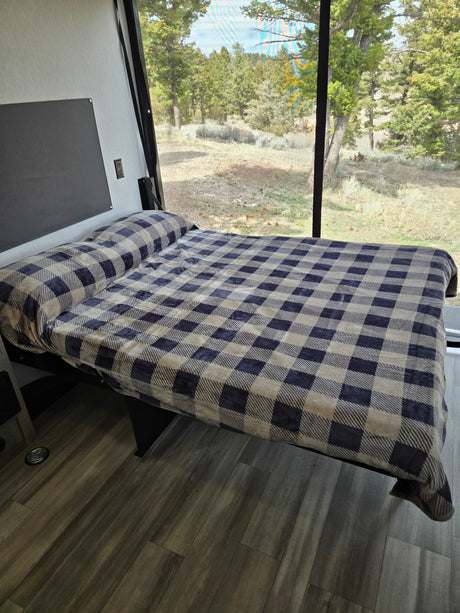 This RV rental features a cozy bed with a blue and beige plaid blanket, positioned by a large window showing a green forest view.