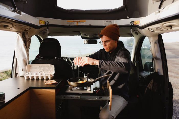 Rv rental adventure: Man cooking breakfast inside a camper van, cracking eggs into a pan on a portable stove by the ocean.