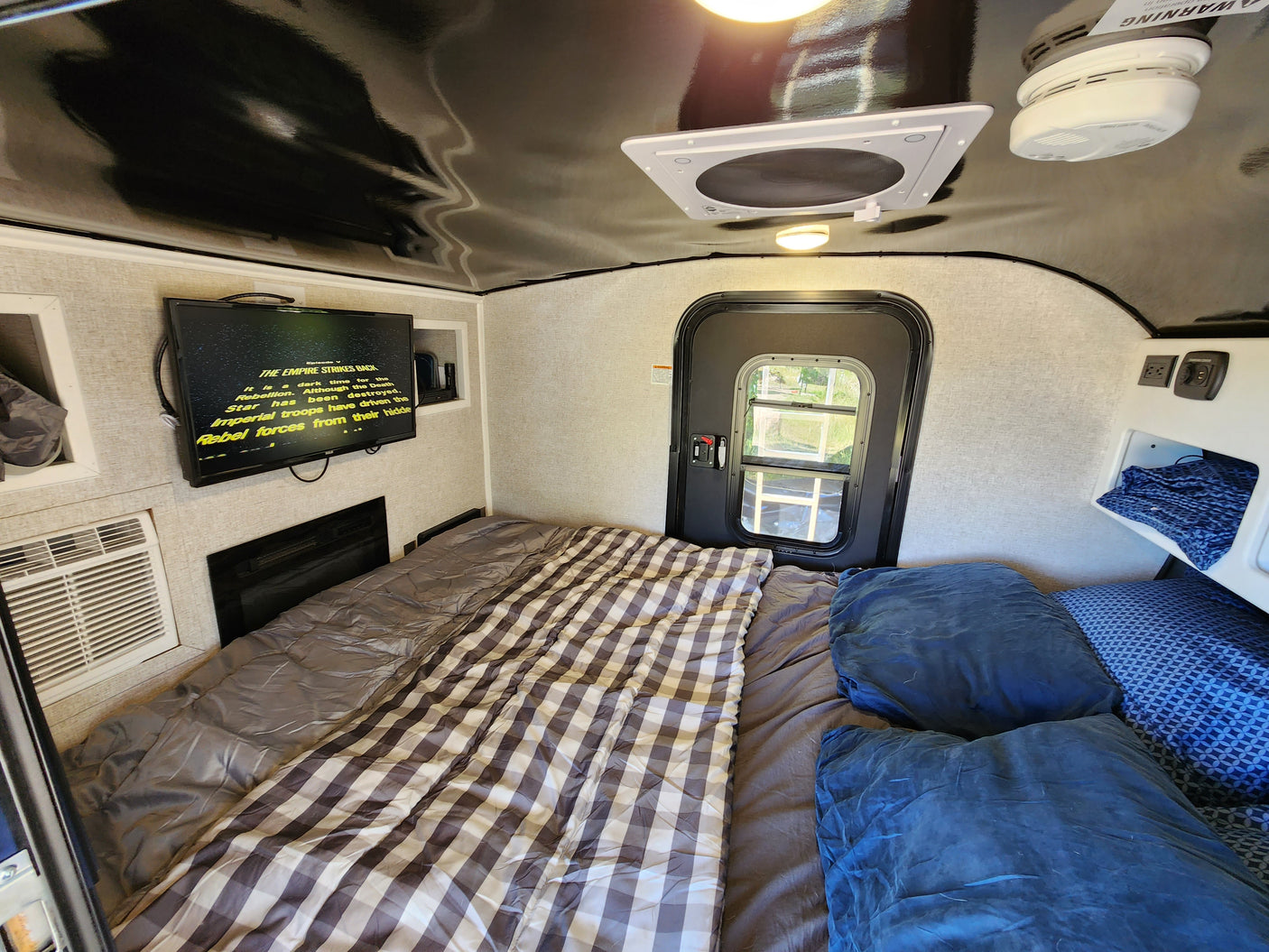 This RV rental features a cozy bed with plaid and gray bedding, a TV playing Star Wars, AC, and a door to nature.