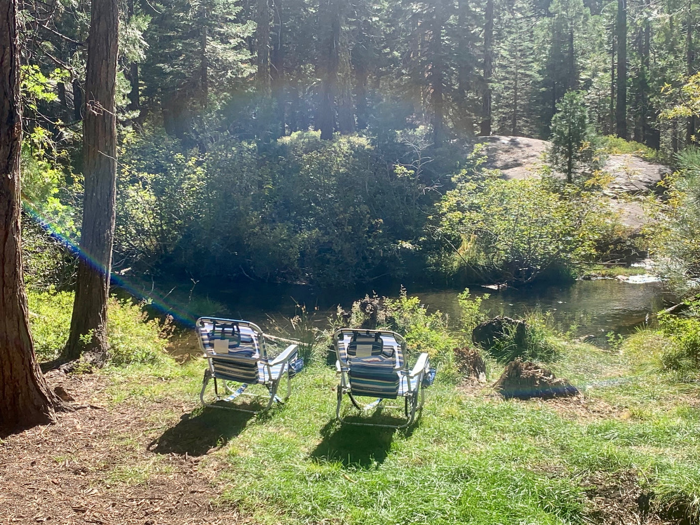 Find peace with your RV rental: Two chairs beside a clear stream in a sunlit forest.