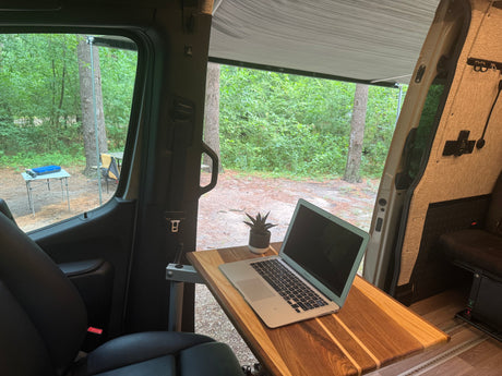 An RV rental offers a perfect mobile office setup with a laptop and plant, looking out at a tranquil campsite in the woods.