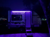 Night view of an RV rental camper with vibrant purple lights illuminating the side, steps, and ground, perfect for an evening setup.