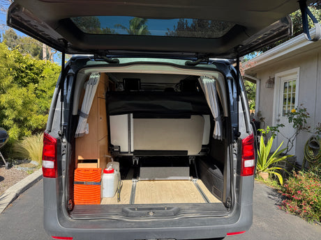 Rear view of an RV rental with tailgate open, showing a folded bed, wooden interior, and travel gear.