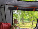 Baby views lush nature from a rooftop tent on an RV rental adventure, with an RV visible outside.