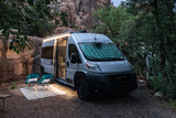 This RV rental camper van, lights on, is set up on a gravel pad with outdoor seating and rug, nestled among rocky cliffs and trees.