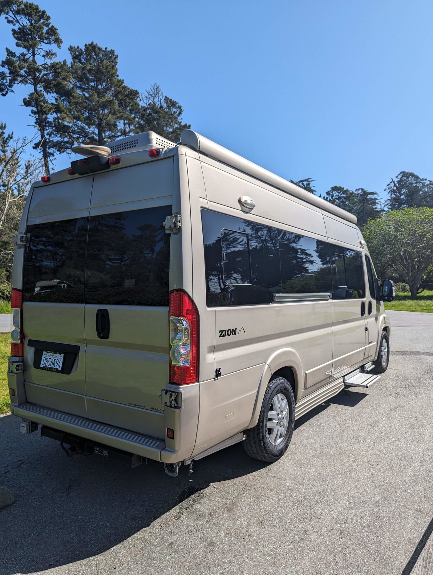 Light-colored Roadtrek Zion camper van, ideal for RV rental. Parked on asphalt with trees & blue sky in background.