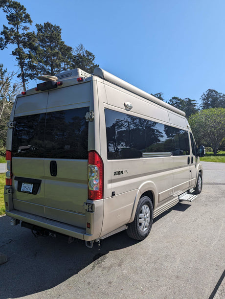 Light-colored Roadtrek Zion camper van, ideal for RV rental. Parked on asphalt with trees & blue sky in background.