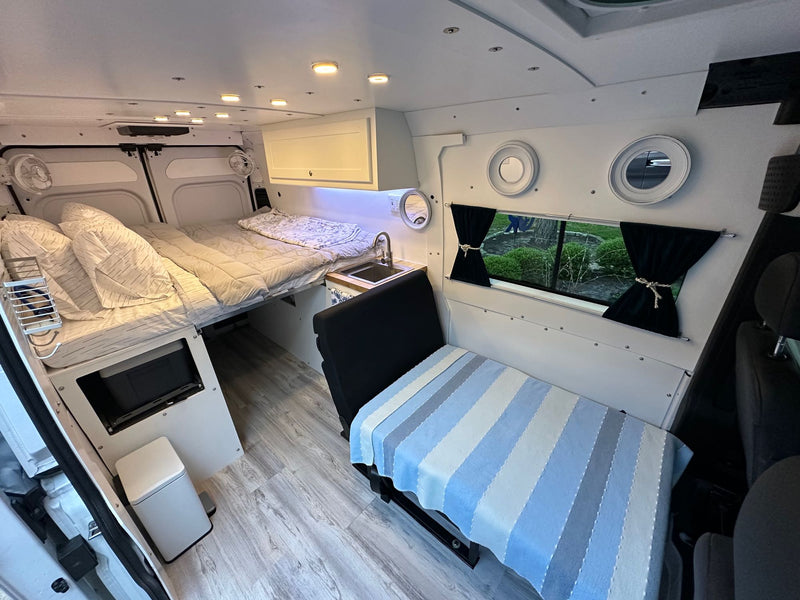  and convertible seating with unique porthole windows.|White RV rental van on a scenic mountain road with snow-capped peaks and autumn trees. Plan your adventure.|Inside an RV rental van: a cozy bed — photo 7