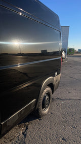 Side view of a black van with rugged tires, reflecting bright sunlight and showing specks on its surface. Great for an RV rental.