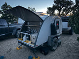 Modern teardrop rv rental camper with an open outdoor kitchen setup, parked on sand alongside a large truck.