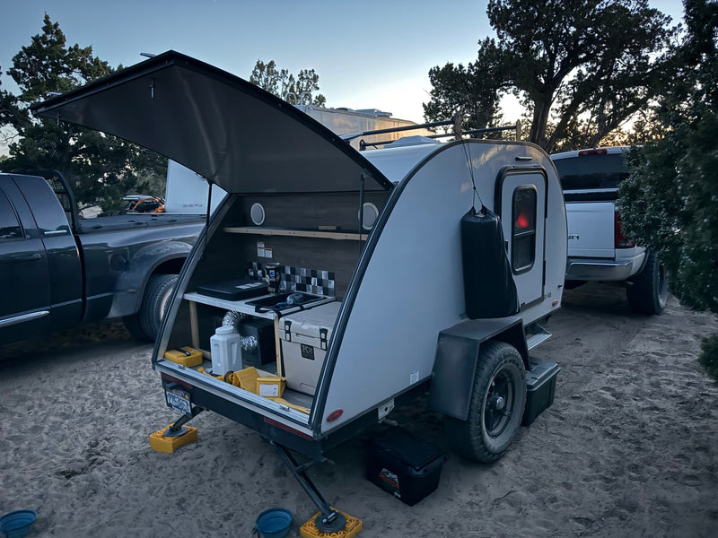 Modern teardrop rv rental camper with an open outdoor kitchen setup, parked on sand alongside a large truck. — photo 8