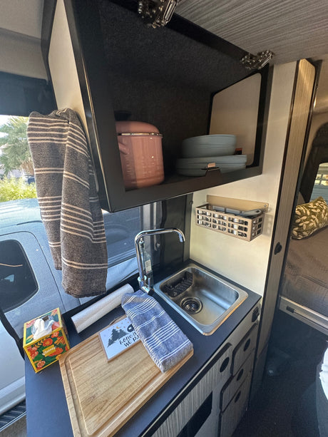 A well-equipped RV rental kitchen features a sink, faucet, overhead cabinet with dishes, spice rack, and counter with cutting board.