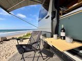 Beachside serenity with an rv rental camper van: awning out, wine & mugs on table, chair ready for ocean views.