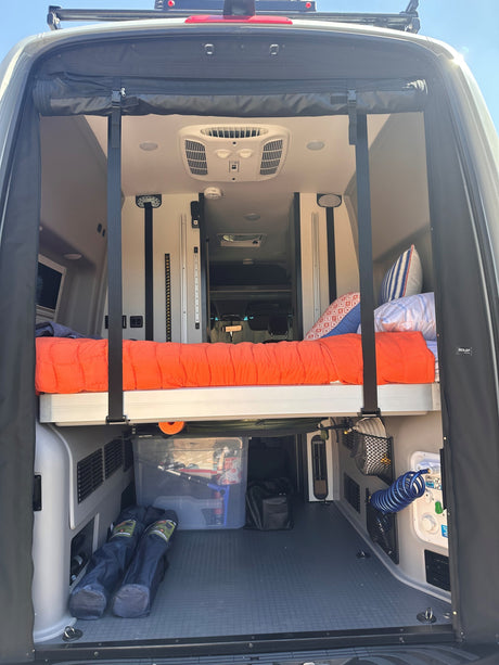 RV rental interior: View from the open rear of a camper van shows an elevated bed, storage, and utility hookups.