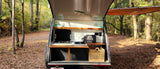 RV rental: Back of a teardrop camper showing a compact kitchen with stove, sink, and wood shelves, set in a peaceful forest.
