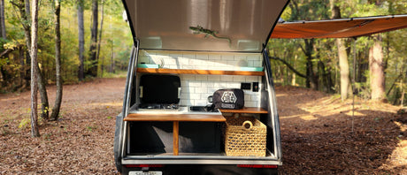 RV rental: Back of a teardrop camper showing a compact kitchen with stove, sink, and wood shelves, set in a peaceful forest.