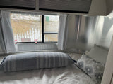 Inside an RV rental: a comfy Airstream bedroom with metallic walls, bed, striped pillow, and window view with emergency exits.