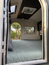 View inside a modern RV rental: a comfy bed, ceiling skylight, and a large arched window revealing a lush outdoor scene.