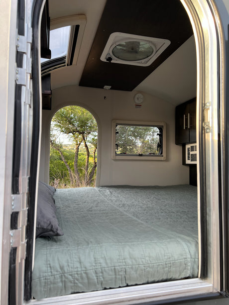 View inside a modern RV rental: a comfy bed, ceiling skylight, and a large arched window revealing a lush outdoor scene.