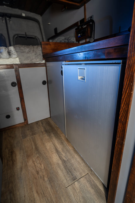An RV rental interior reveals a built-in silver fridge, wooden floor, and gray storage cabinets.