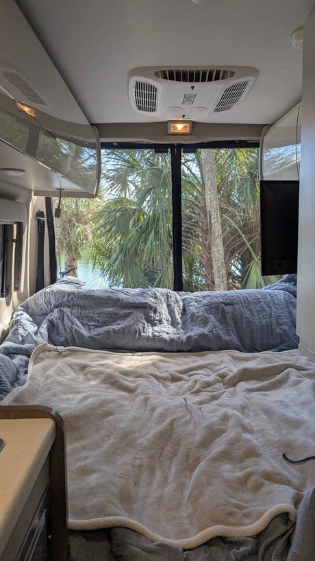 Cozy RV rental bed with a tropical view of palm trees and water, seen from inside the camper van's comfortable interior.