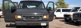 Front view of a dark grey Ford van with open doors in a lot with other rental vans. Great for an rv rental trip.