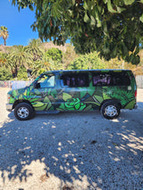 This vibrant green RV rental van, custom-painted with leaf and rose designs, is parked on gravel under a tree on a sunny day.