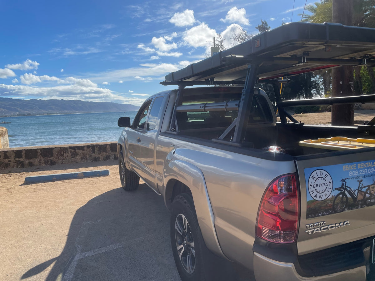 A silver Toyota Tacoma pickup with a bed rack, parked by the ocean with mountains. Great for e-bike & RV rentals in Hawaii. — photo 4