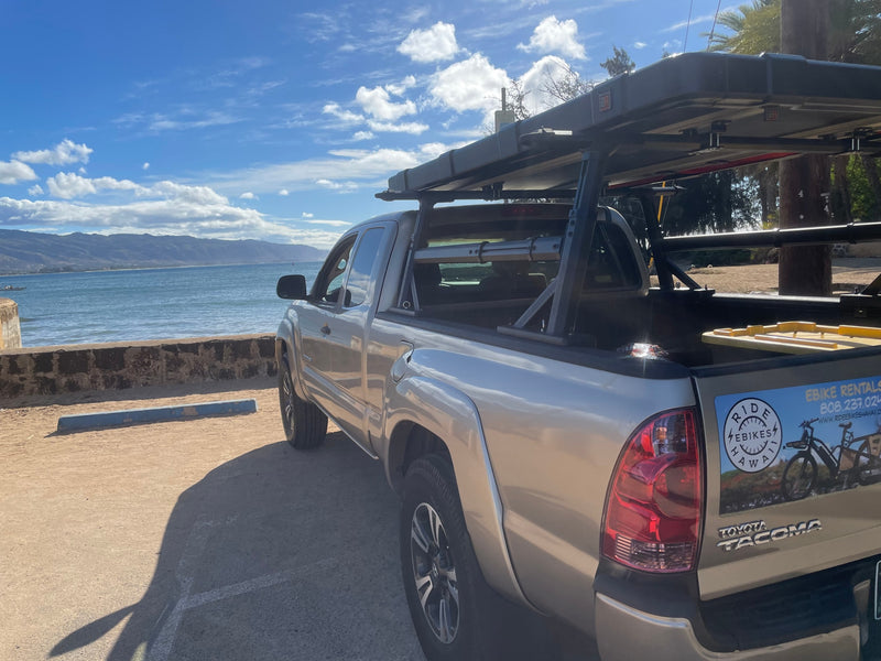 A silver Toyota Tacoma pickup with a bed rack, parked by the ocean with mountains. Great for e-bike & RV rentals in Hawaii. — photo 4