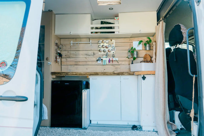  and storage solutions.|Hands stir a vibrant vegetable stir-fry on a portable stove. Ideal outdoor cooking setup for your RV rental trip!|RV rental adventure: Two people and their dogs prepare food outside a white camper van in a mountain forest setting.|A bright RV rental interior featuring a compact van conversion kitchen with wood accents