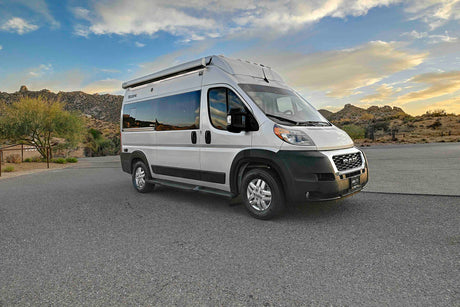 White camper van RV rental parked on asphalt, ready for a desert road trip among rocky mountains and a dramatic sky.