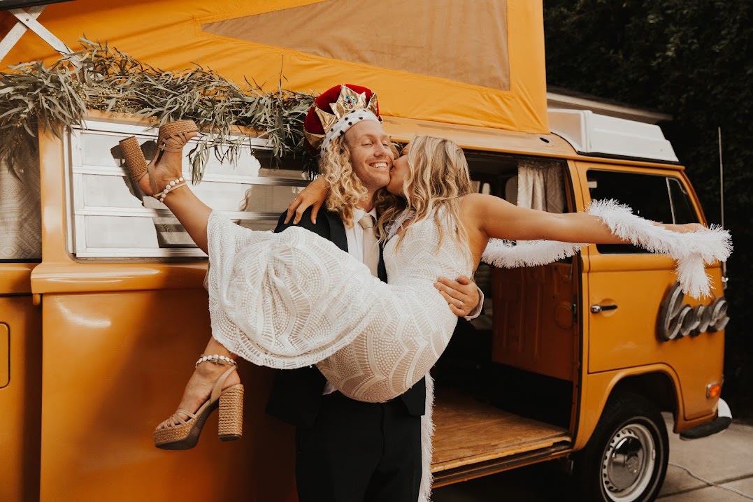 A joyful couple celebrates by a vintage orange RV rental. He wears a crown, she a white dress, as he lifts her for a kiss, embodying fun and romance. — photo 14