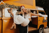 A joyful couple celebrates by a vintage orange RV rental. He wears a crown, she a white dress, as he lifts her for a kiss, embodying fun and romance.