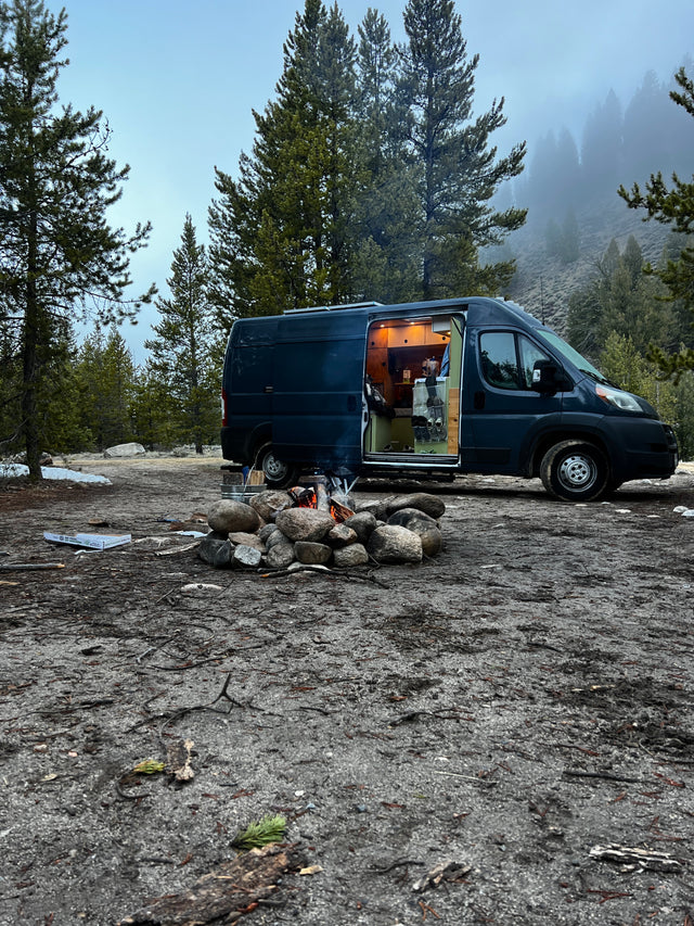 Cozy RV rental camping scene: dark blue camper van with illuminated interior, warm campfire, misty forest backdrop.
