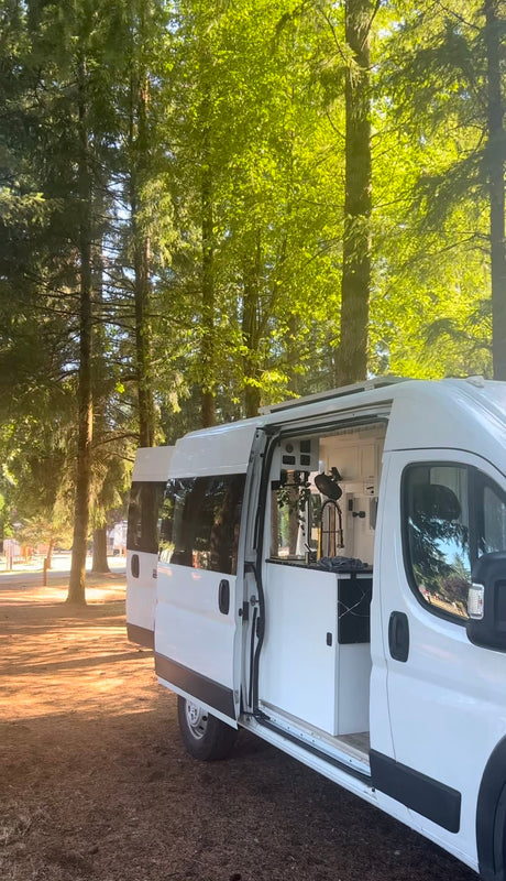 A bright white RV rental with its side door open, revealing a modern custom interior kitchen, parked among tall trees for camping.